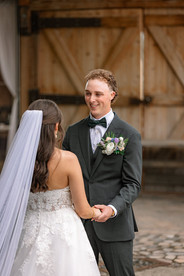 Camera facing the groom as he holds hands with the bride during an outdoor ceremony, her back to the lens and veil flowing behind her as they stand facing each other at one of the romantic Southern Alberta Wedding Venues.