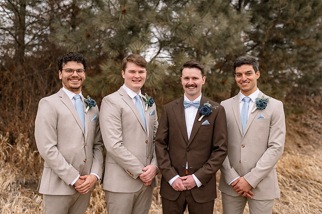 Groom and groomsmen posing together outdoors at an Airbnb wedding venue, wearing light grey suits and blue accents, with trees and natural scenery in the background.