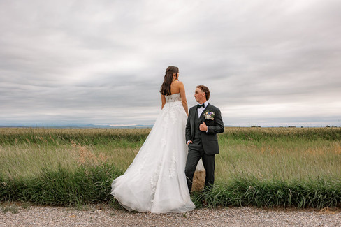 Bride in a long white gown standing in a grassy field with the groom beside her, captured by a Southern Alberta wedding photographer.