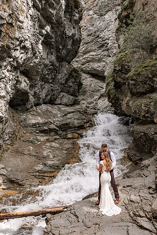 Bride standing alone beside cascading waterfall surrounded by rugged rock walls on their Crowsnest Pass wedding day 