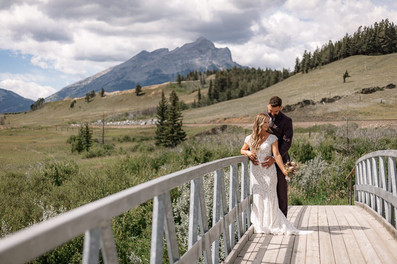 Bride and groom standing on a bridge with mountains and open fields in the background on their Crowsnest Pass wedding day