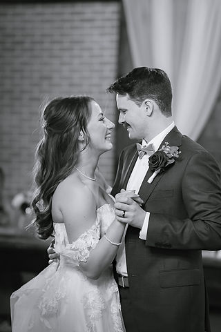 Black-and-white portrait of the bride and groom sharing their first dance and smiling at one another during their wedding reception, captured by Olds wedding photographers.