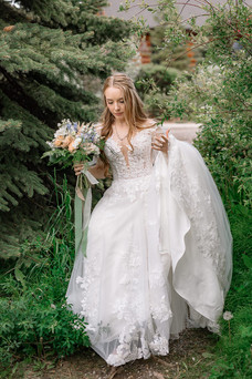 Bride holding her bouquet and walking through the trees during her Canmore Nordic Centre wedding, with her flowing wedding dress and mountain surroundings creating a romantic portrait.