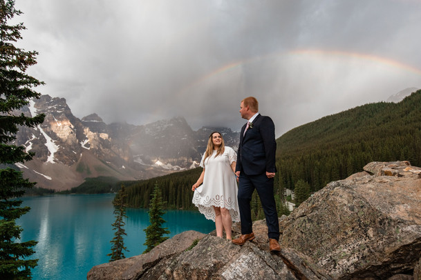 Bride and groom standing on the rocks. Bride is holding out the bottom of her dress. Rainbow in the sky behind them, the lake and mountains.