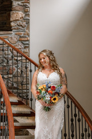 Bride holding a colorful wildflower bouquet while standing on a curved staircase inside a rustic farmhouse during her romantic farm wedding.