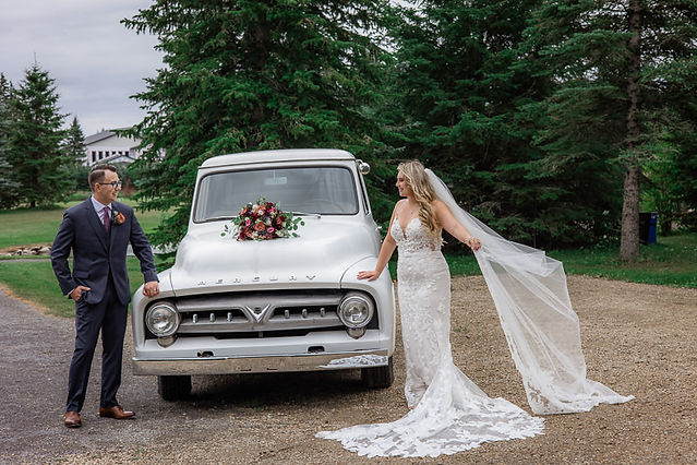 bride smiles at groom looking across front of truck with bouquet on top