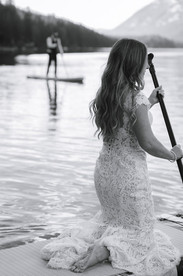 Bride sitting on a paddleboard through shallow lake water while the groom stands on his paddleboard in the distance on their Crowsnest Pass wedding day