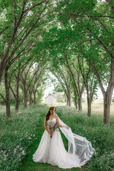 Bride standing alone beneath a tunnel of trees, her long veil flowing behind her as she holds a bouquet on a lush green path lined with wildflowers at one of the stunning Southern Alberta Wedding Venues.