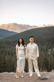 The bride and groom stand side by side on a rocky overlook, dressed in traditional chinese outfits, with a sweeping forest and soft mountain skyline behind them at sunset