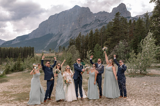 A Canmore wedding photographer captures the bride, groom, bridesmaids, and groomsmen celebrating with champagne and raised bouquets against the stunning Rocky Mountain backdrop after the ceremony.