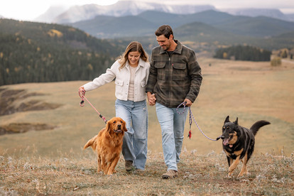 Couple walking with their dogs through an open field with scenic mountain views during fun engagement ideas.
