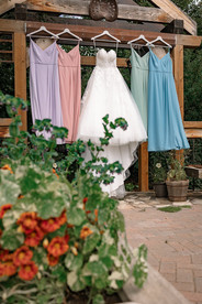 White wedding dress and pastel bridesmaids’ dresses hanging from a wooden outdoor structure, with greenery and floral arrangements in the foreground at a Coutts Centre wedding.