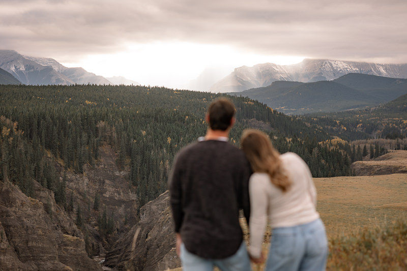 Couple holding hands and overlooking a mountain landscape during their Sheep River engagement photos, with forested cliffs and moody skies in the background.