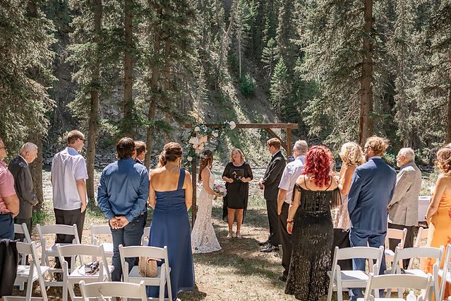 Wide view of an outdoor wedding ceremony with guests standing on either side as the couple exchanges vows beneath a floral arch with the officiant at  Cochrane wedding venues