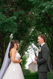 Bride and groom facing each other beneath tall trees during their outdoor ceremony, exchanging vows and smiling with a blurred house and trees in the background at one of the scenic Southern Alberta Wedding Venues.