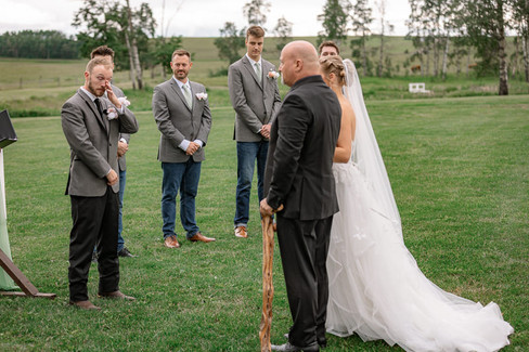 At Diamond in teh Rough, the bride stands beside her father at the alter, facing the groom while the groom wipes his tears during the outdoor ceremony on the green lawn