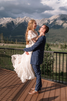 A Canmore wedding photographer captures the groom lifting the bride on a scenic mountain view deck, with dramatic Rocky Mountain peaks and soft evening light creating a romantic wedding portrait.