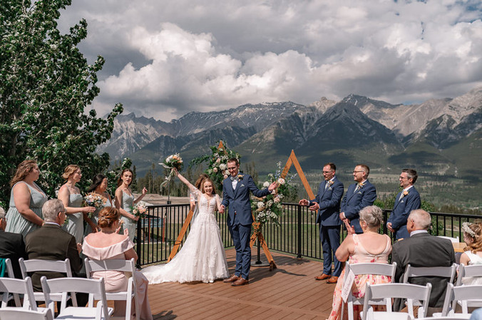 Bride and groom celebrate at one of the most scenic Canmore wedding venues, walking back down the aisle after their ceremony beneath a floral arch with their wedding party, guests, and the Rocky Mountains in the background.
