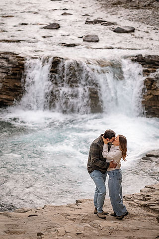 Couple kissing on a rocky riverbank in front of a waterfall during Sheep River engagement photos, with rushing water and natural stone creating a romantic, adventurous setting.