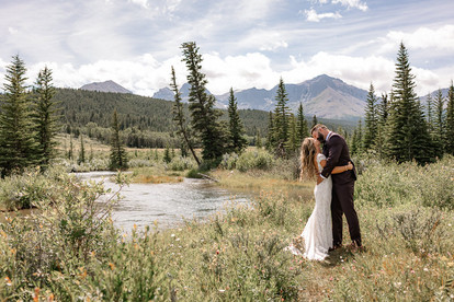 Bride and groom standing together beside a river surrounded by evergreen trees and mountain views on their Crowsnest Pass wedding day
