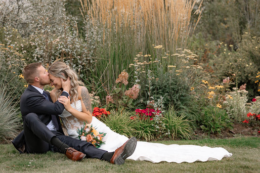 Bride and groom sitting on the grass at The Sensory Canmore, sharing a kiss surrounded by lush garden flowers.