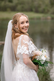 Bride smiling over her shoulder while holding her bouquet during her Canmore Nordic Centre wedding, with a peaceful mountain lake creating a romantic Rocky Mountain backdrop.