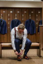 Groom tying up shoes with groomsmen jackets hanging in the background