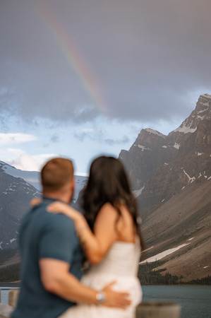 A couple stands close together with their backs to the camera as a rainbow arcs across the cloudy sky above the mountains, creating a peaceful and romantic moment in the Rockies.