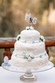Three-tier white wedding cake displayed outdoors on a wooden deck at a Bragg Creek wedding venue.