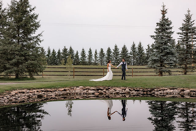 Bride and groom walking in front of pond holding hands, reflection of bride and groom in the pond