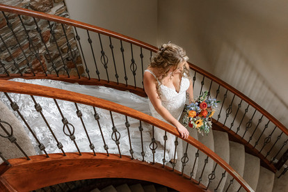 Bride holding a colorful bouquet while walking down a curved staircase inside a rustic farmhouse during her romantic farm wedding preparations.