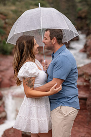 Under an umbrella, a couple in a white dress and blue shirt smiles joyfully together during their engagement photoshoot in Waterton.