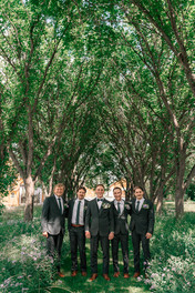 Groom and his four groomsmen in matching suits standing together beneath leafy trees outdoors at a Coutts Centre wedding.