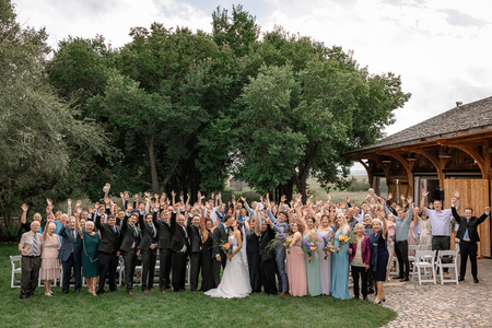Wide shot of a large group of wedding guests gathered around the bride and groom as they share a kiss, guests cheering with raised arms beside a barn and a tree-lined ceremony space at one of the vibrant Southern Alberta Wedding Venues.