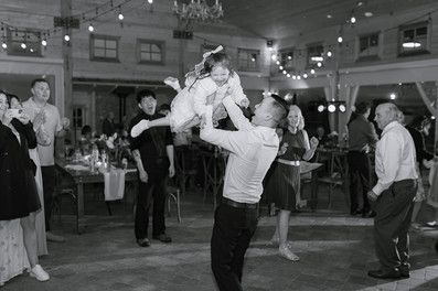 Guests celebrating at a wedding reception in black and white, with a man lifting a child into the air, captured by a Southern Alberta wedding photographer.