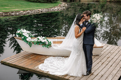 Bride ad groom standing on dock with white canoe behind them that is decorated with white and green florals