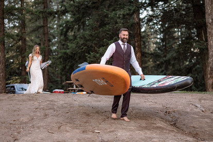 Groom carrying an inflatable paddleboard near a forest campsite on their Crowsnest Pass wedding day