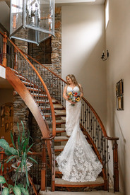 Bride holding a colorful bouquet while posing on a grand curved staircase inside a rustic farmhouse during her romantic farm wedding preparations.