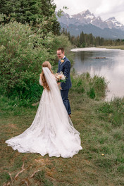 Bride and groom sharing a quiet moment during their Canmore Nordic Centre wedding, with a tranquil mountain lake, evergreen forest, and dramatic Rocky Mountain peaks in the background.