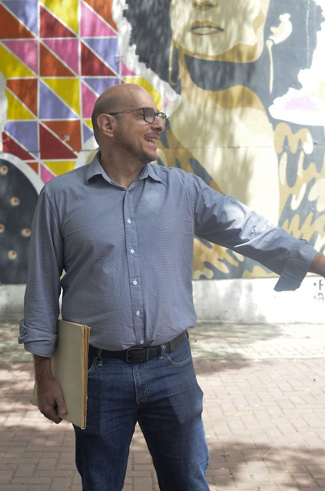 Gonzalo segurando jornais antigos na frente de pinturas da UFCG sorrindo para o lado direito da foto
