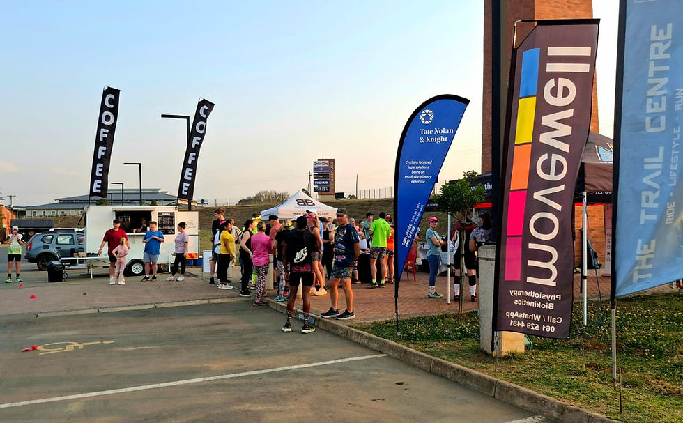 Runners gather at a lively outdoor race start beside COFFEE banners, a coffee trailer, and sponsor flags at sunset.