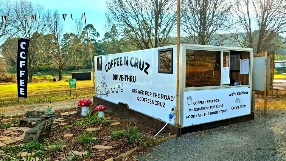 Coffee N Cruz drive-thru kiosk in a park, with COFFEE sign, cashless cards-only notice, and mushroom decor.