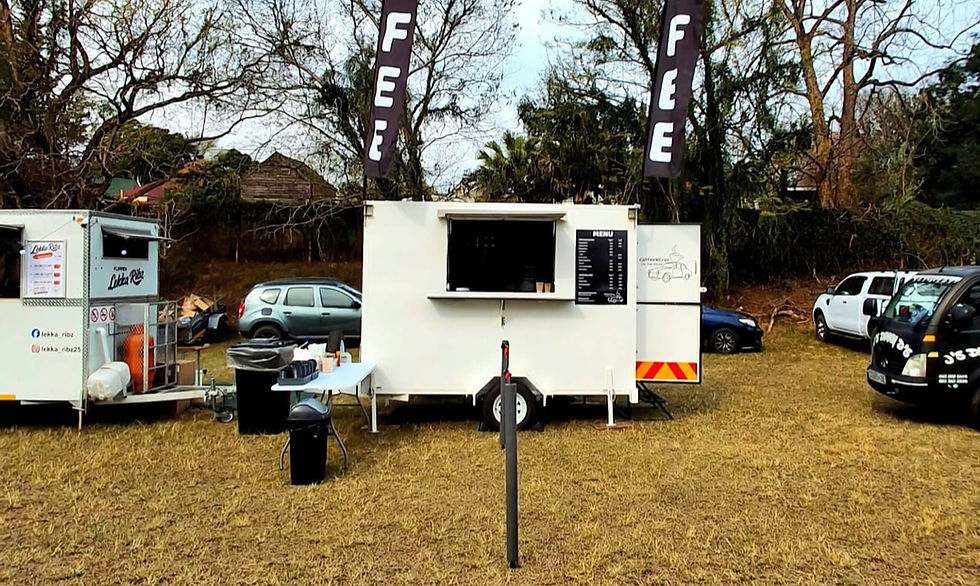 Outdoor coffee trucks and a white menu trailer with FEE banners, parked on grass among cars and trees.