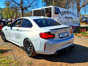 White BMW coupe at Coffee n Cruz drive-thru during a sunny car meet, with people gathered and a coffee trailer behind.