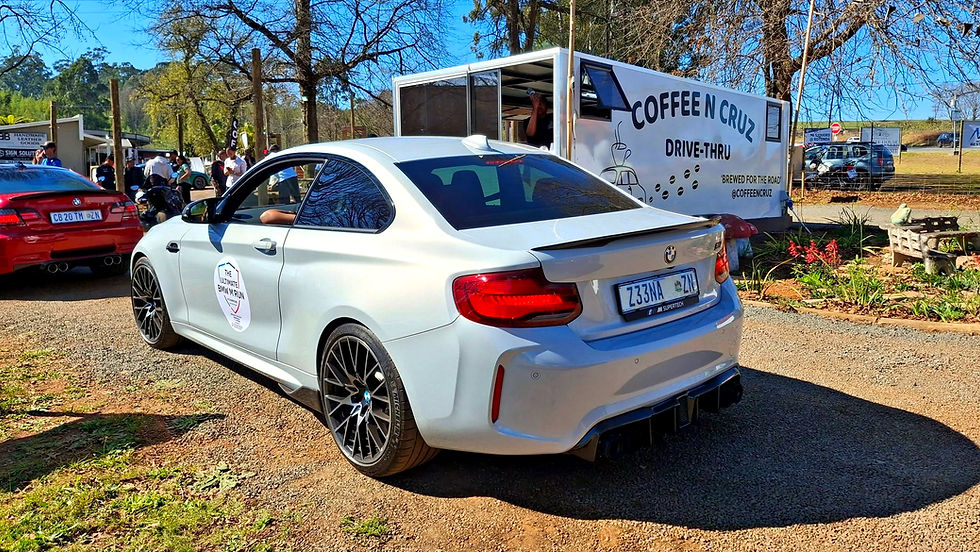 White BMW coupe at Coffee n Cruz drive-thru during a sunny car meet, with people gathered and a coffee trailer behind.