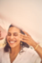 A woman smiles with closed eyes, touching her hair. She wears a white shirt, gold jewelry, and stands in a soft, pastel background.
