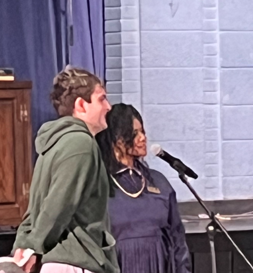 A white young man wearing a hoodie and an African American woman in a dress stand in front of a microphone.