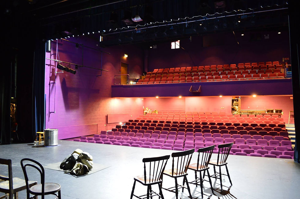 View from a theatre stage toward rows of auditorium seats, with chairs and rehearsal props in the foreground.