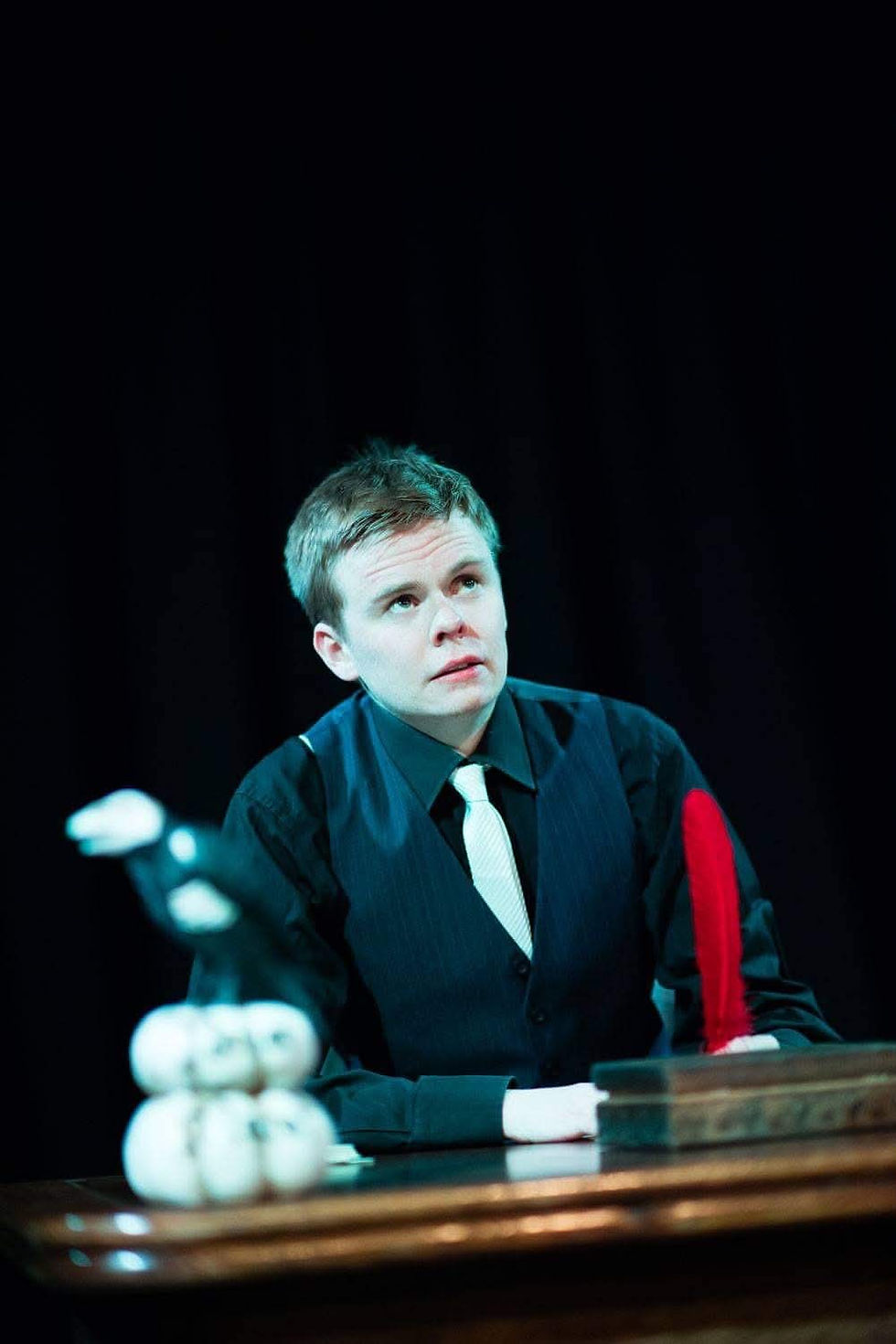 Actor in formal costume seated at a desk, looking upward under theatrical lighting.