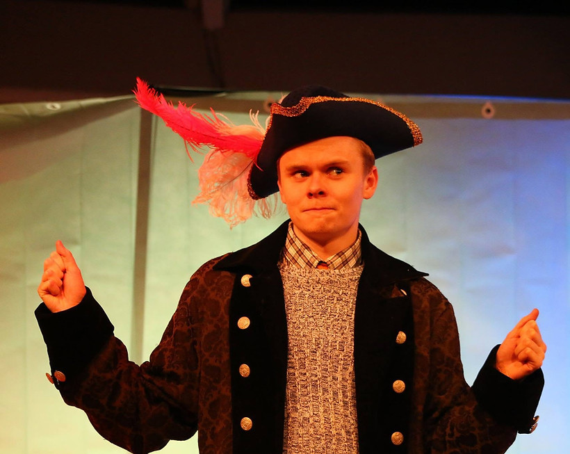 Michael Fish performing on stage in period costume and wearing a feathered hat.